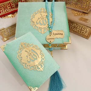 May include: A set of three decorative boxes with gold trim and a green box with a gold inscription of the word "Allah" in Arabic. The boxes are covered in a soft, velvety fabric. A blue tassel hangs from the green box. A blue beaded necklace with a gold pendant that reads "Lyanna" is included.