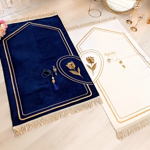 May include: Two prayer rugs with gold trim and a floral design. One rug is navy blue and the other is white. The white rug has the name "Ayesha" embroidered on it.
