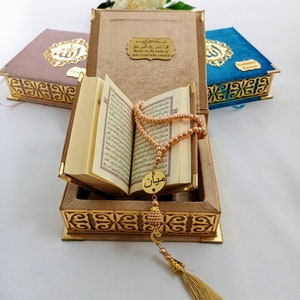 May include: An open Quran with a gold-colored tassel and prayer beads resting on top of the book. The book is in a brown velvet box with gold-colored trim and an inscription in Arabic that reads "Recite in the name of your Lord who created".