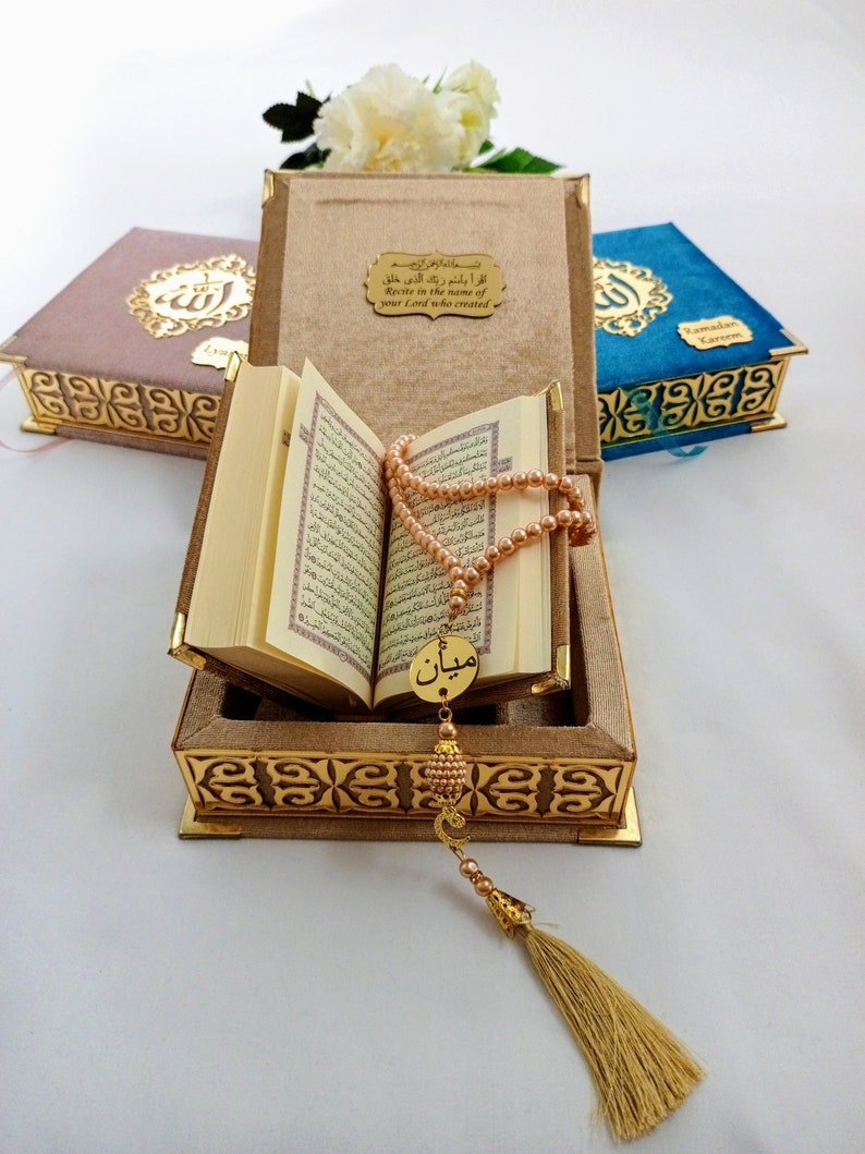 May include: An open Quran with a gold-colored tassel and prayer beads resting on top of the book. The book is in a brown velvet box with gold-colored trim and an inscription in Arabic that reads "Recite in the name of your Lord who created".