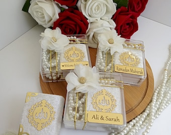 May include: Three clear boxes with white velvet interiors and gold trim contain white pearl prayer beads. Each box has a gold decorative design with the Arabic word for God. The boxes are on a wooden surface with white roses and red roses in the background. The boxes have personalized messages: "Welcome Baby", "Ramadan Mubarak", and "Ali & Sarah".