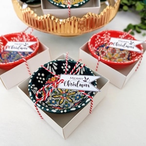 May include: Four small ceramic bowls with floral patterns, two red and two green, are packaged in white boxes with red and white twine and a "Merry Christmas" tag.