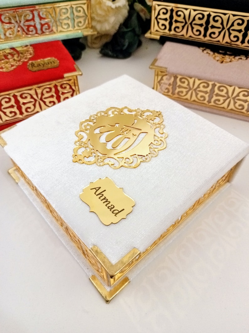 May include: A white velvet box with gold trim and a gold Arabic calligraphy design. The box has a gold nameplate that says "Ahmad".