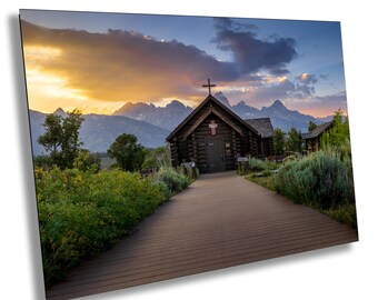 May include: A wooden chapel with a cross on the roof sits in front of a mountain range at sunset. The chapel has a wooden walkway leading up to the entrance. The sky is a mix of blue, purple, and orange.