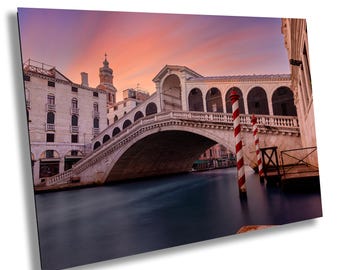 Puede incluir: Una vista de un puente sobre un canal en Venecia, Italia. El puente está hecho de piedra y tiene un arco redondeado. El canal está tranquilo y refleja el cielo, que es de un rosa y morado suave. El puente está iluminado por el sol poniente, proyectando largas sombras.
