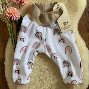 May include: White baby pants with a brown waistband and brown cuffs. The pants have a rainbow pattern with pink, brown, and gray colors. The pants have a wooden tag attached with the text "Nähwerkstatt" and "Handgemacht".