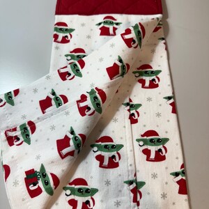 May include: White kitchen towel featuring a repeating pattern of a green alien character in a Santa hat and red outfit. The towel has a red quilted top with a white button.