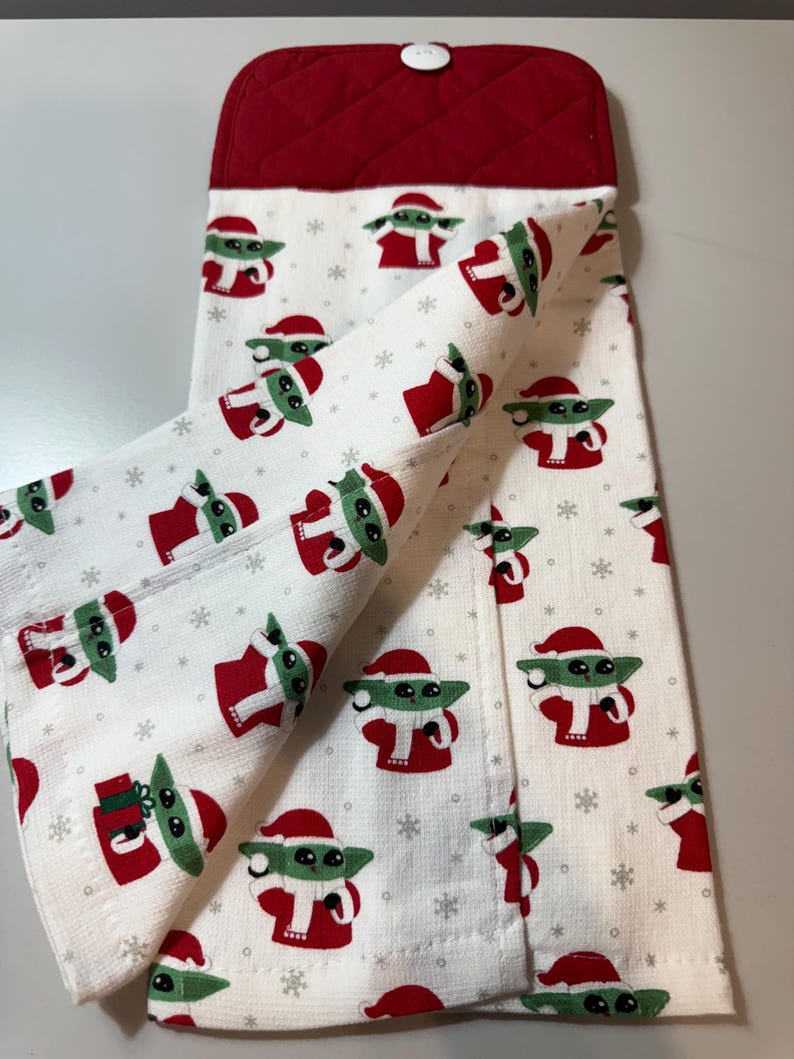 May include: White kitchen towel featuring a repeating pattern of a green alien character in a Santa hat and red outfit. The towel has a red quilted top with a white button.