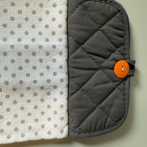 May include: A kitchen towel with a gray and white polka dot pattern on one side and a quilted gray fabric on the other. An orange button secures the towel. The towel is designed to hang.