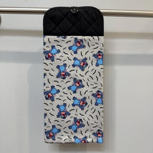 Puede incluir: Un pa&ntilde;o de cocina colgante con una parte superior acolchada negra y una parte inferior estampada. La parte inferior presenta un fondo gris claro con personajes de dibujos animados azules y formas negras de murci&eacute;lagos. El pa&ntilde;o cuelga de una barra plateada.