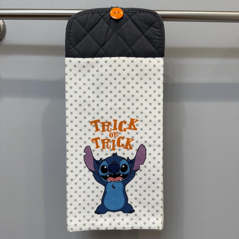 May include: A hanging kitchen towel with a black quilted top and a white dotted body. The towel features a blue cartoon character and the words "Trick or Treat" in orange. An orange button secures the top.