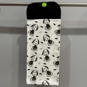 May include: A hanging kitchen towel with a black top and a green button. The towel features a repeating pattern of Snoopy dressed as a skeleton, along with black bat shapes, on a white background.