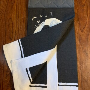 May include: Black and white kitchen towel with a graphic of a crescent moon and bats. The towel has a white border with black stripes. The towel is folded in half and has a gray oven mitt attached.