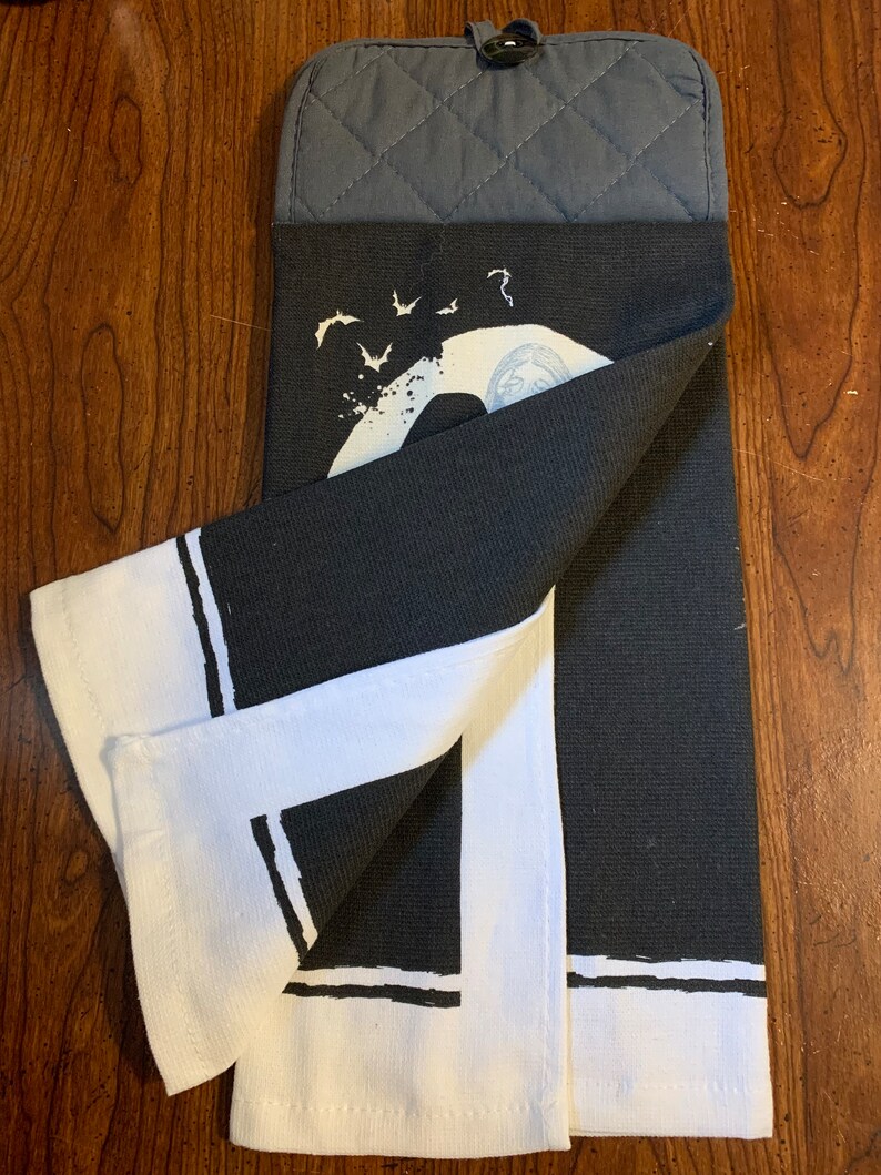 May include: Black and white kitchen towel with a graphic of a crescent moon and bats. The towel has a white border with black stripes. The towel is folded in half and has a gray oven mitt attached.