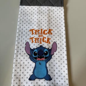 May include: A white kitchen towel with gray polka dots and a gray quilted top. The towel features a blue cartoon character and the orange text "Trick or Treat".