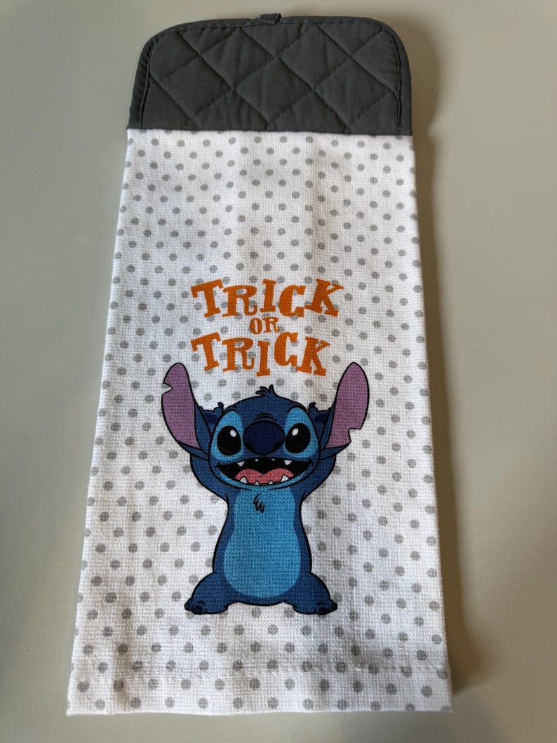May include: A white kitchen towel with gray polka dots and a gray quilted top. The towel features a blue cartoon character and the orange text "Trick or Treat".