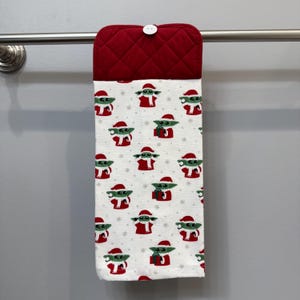 May include: A hanging kitchen towel with a red quilted top and a white button. The towel features a repeating pattern of a green character wearing a Santa hat and red outfit, holding a gift. The background is white with small snowflakes.