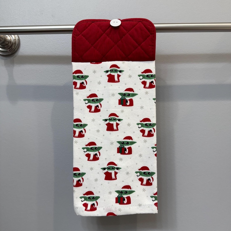 May include: A hanging kitchen towel with a red quilted top and a white button. The towel features a repeating pattern of a green character wearing a Santa hat and red outfit, holding a gift. The background is white with small snowflakes.