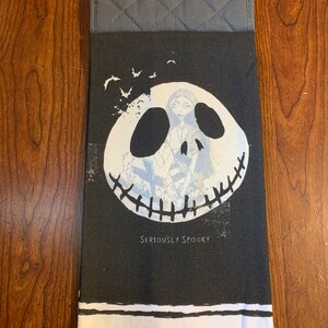 May include: Black and white kitchen towel with a graphic of Jack Skellington from The Nightmare Before Christmas. The towel has a gray quilted potholder attached to the top. The text "Seriously Spooky" is printed on the towel.