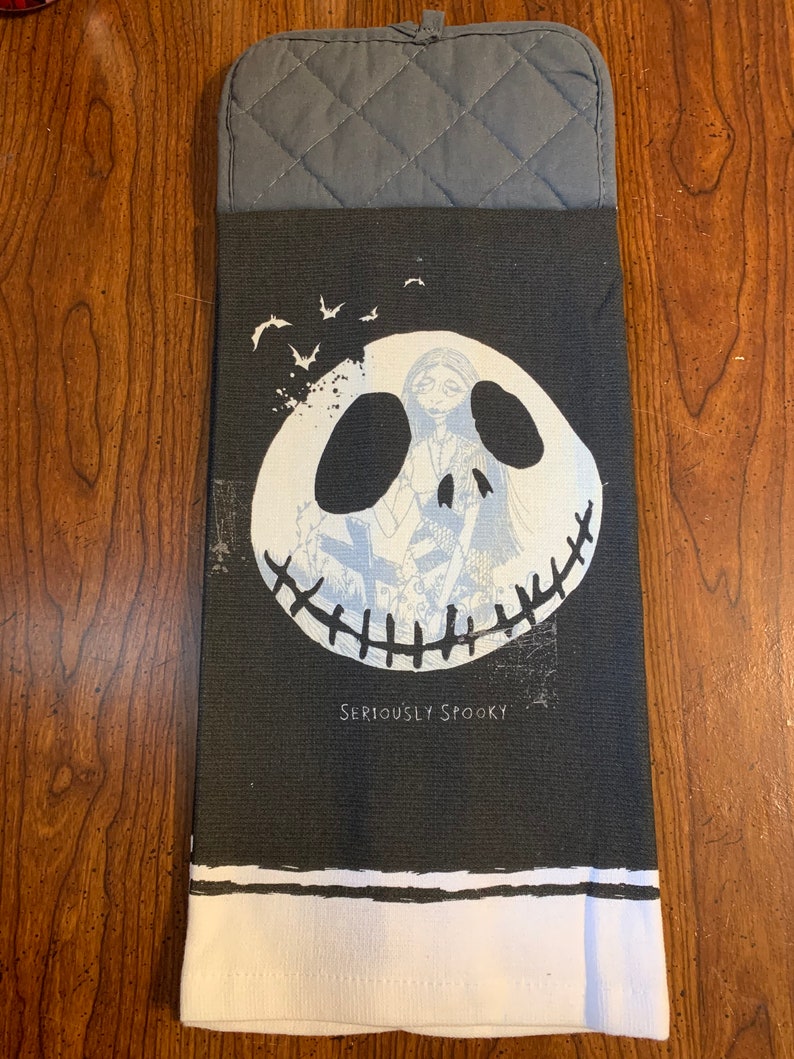 May include: Black and white kitchen towel with a graphic of Jack Skellington from The Nightmare Before Christmas. The towel has a gray quilted potholder attached to the top. The text "Seriously Spooky" is printed on the towel.