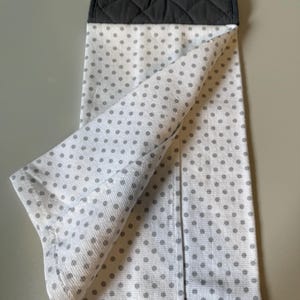 May include: White kitchen towel with gray polka dots, featuring a quilted dark gray top section. An orange button secures the towel. The towel is folded, showcasing the polka dot pattern.