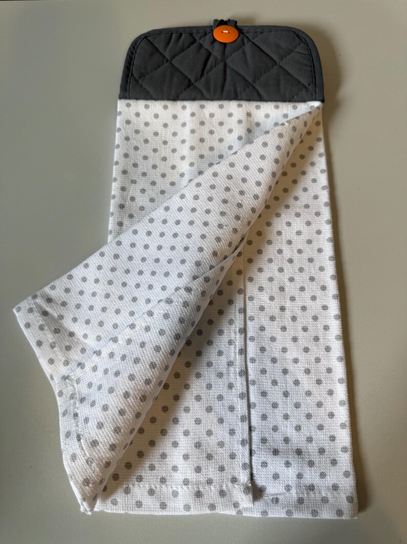 May include: White kitchen towel with gray polka dots, featuring a quilted dark gray top section. An orange button secures the towel. The towel is folded, showcasing the polka dot pattern.