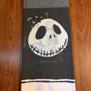 May include: Black and white kitchen towel with a gray potholder. The towel features a black and white illustration of Jack Skellington's face from the Nightmare Before Christmas movie. The text "Seriously Spooky" is printed below the illustration.