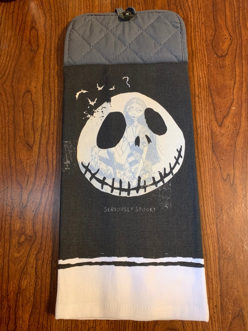 May include: Black and white kitchen towel with a gray potholder. The towel features a black and white illustration of Jack Skellington's face from the Nightmare Before Christmas movie. The text "Seriously Spooky" is printed below the illustration.