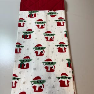 May include: A white kitchen towel with a red quilted top. The towel is patterned with green and white cartoon characters wearing Santa hats and red outfits, with snowflakes and gift boxes. The towel is designed for the Christmas holiday season.
