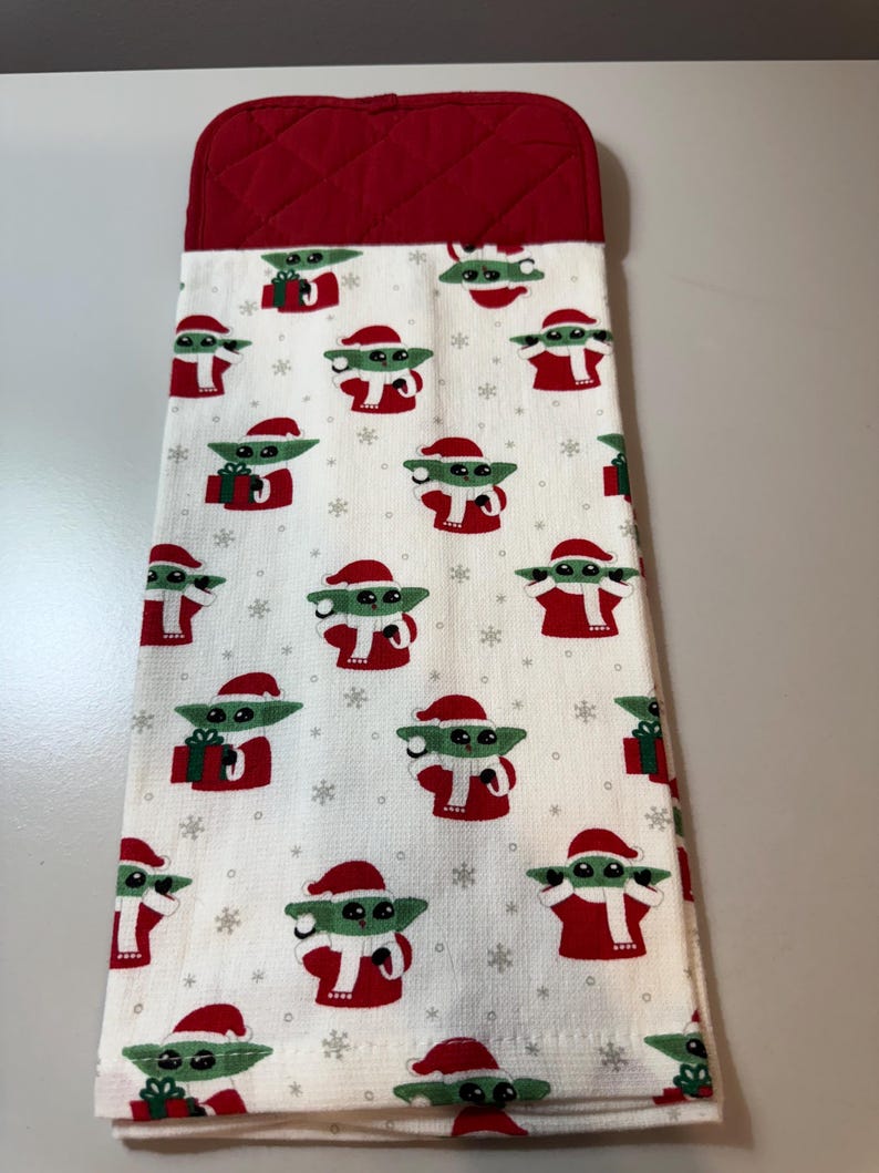 May include: A white kitchen towel with a red quilted top. The towel is patterned with green and white cartoon characters wearing Santa hats and red outfits, with snowflakes and gift boxes. The towel is designed for the Christmas holiday season.