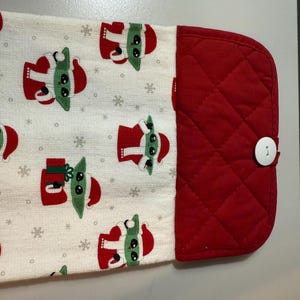 May include: A white kitchen towel featuring a repeating pattern of a green-skinned character in a red Santa suit and snowflakes. The towel has a red quilted section with a white button.