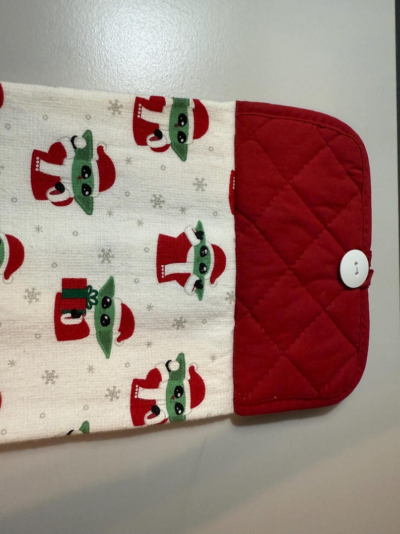 May include: A white kitchen towel featuring a repeating pattern of a green-skinned character in a red Santa suit and snowflakes. The towel has a red quilted section with a white button.