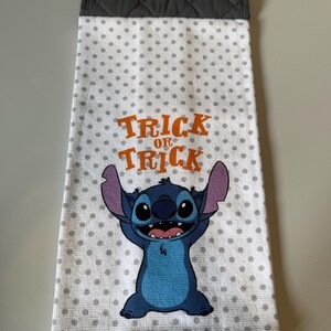 May include: A white kitchen towel with gray polka dots and a cartoon character. The towel has the words "Trick or Treat" in orange. The top of the towel is gray with a quilted pattern and an orange button.