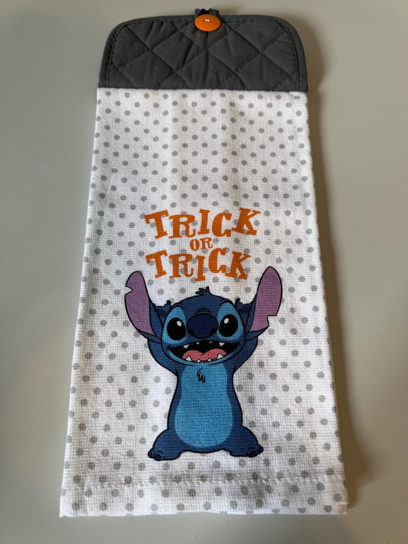 May include: A white kitchen towel with gray polka dots and a cartoon character. The towel has the words "Trick or Treat" in orange. The top of the towel is gray with a quilted pattern and an orange button.