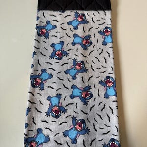 Puede incluir: Un pa&ntilde;o de cocina con un estampado del personaje Stitch de Lilo & Stitch. El pa&ntilde;o tiene un fondo gris claro con figuras azules de Stitch y dise&ntilde;os negros en forma de murci&eacute;lago. La parte superior es negra y acolchada.