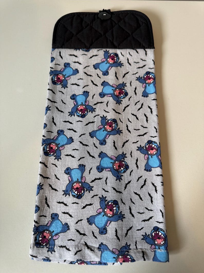 Puede incluir: Un pa&ntilde;o de cocina con un estampado del personaje Stitch de Lilo & Stitch. El pa&ntilde;o tiene un fondo gris claro con figuras azules de Stitch y dise&ntilde;os negros en forma de murci&eacute;lago. La parte superior es negra y acolchada.