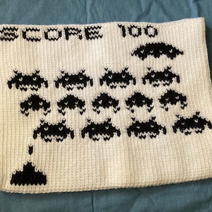 May include: White crocheted pillow with a black pixelated design of a classic video game. The design includes the text "SCORE 100" and multiple spaceships.