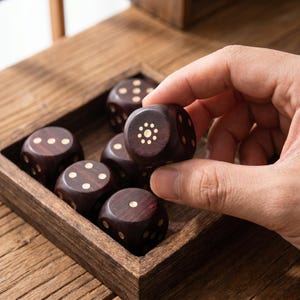 May include: A set of dark brown wooden dice with white dots, displayed in a wooden tray. One die is being held by a hand. The dice are a classic game accessory.