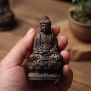 May include: A small, dark brown wooden Buddha figurine carved in a seated meditation pose on a lotus base. The figure features detailed hair texture, draped robes, and a small swastika symbol engraved on the chest area, held in a palm.