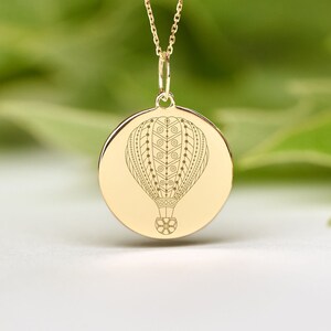 May include: A gold necklace with a round pendant featuring a detailed engraved design of a hot air balloon.