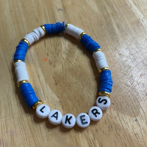 May include: A white and blue beaded bracelet with gold accents. The bracelet spells out "LAKERS" in black letters on white beads.