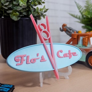 Può includere: Un cartello stampato in 3D blu e rosa con la scritta "Flo's Cafe" e un design stilizzato "V" rosso e bianco. Il cartello è su un supporto bianco e sullo sfondo c'è un camion giocattolo.