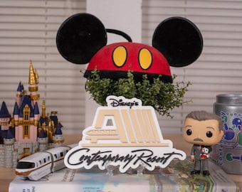 May include: A display of Disney merchandise, featuring a Mickey Mouse ear hat, a Cinderella castle ornament, a Contemporary Resort sign, a toy monorail, a Funko Pop figure, and a silver mug with a handle.