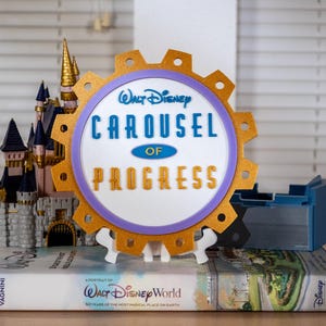 Op de afbeelding: Een decoratief bord met de tekst "Walt Disney Carousel of Progress" in blauw en goud, tegen een witte achtergrond. Het bord is tandwielvormig met een paarse rand en gouden accenten. Een miniatuurkasteel en een boek zijn ook zichtbaar.