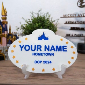 Könnte beinhalten: Weißer ovaler Namensschild mit goldenen Sternen um den Rand. Das Schild hat eine blaue Silhouette von Schloss Aschenputtel, den Text "YOUR NAME", "HOMETOWN" und "DCP 2024".