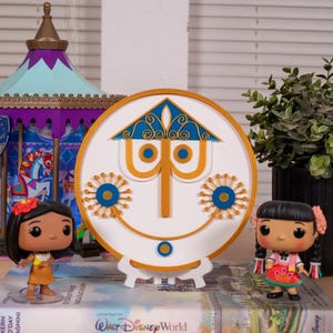 Puede incluir: Una exhibición con una pieza de arte mural circular blanca y dorada con un diseño de cara sonriente, flanqueada por dos figuras de personajes y un carrusel decorativo. Una planta en maceta se encuentra a la derecha.