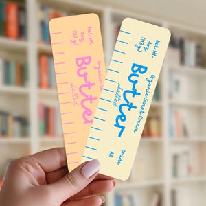 May include: Two rectangular bookmarks designed to resemble sticks of butter. The left bookmark is peach-colored with pink text reading Butter, Salted, and Net Wt 4oz (113g). The right bookmark is cream-colored with blue text reading Butter, Salted, Organic Sweet Cream, and Net Wt 4oz (113g).