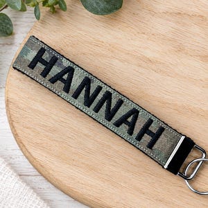 May include: A fabric keychain strap featuring a camouflage pattern in shades of green, brown, and black. The name HANNAH is embroidered in bold black capital letters across the center. A silver metal clasp and a circular split ring are attached.