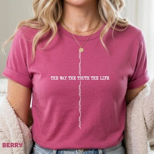 Puede incluir: Una camiseta de manga corta color baya con un diseño gráfico blanco de una cruz formada por la palabra JESUS repetida verticalmente. El texto horizontal a través de la cruz dice THE WAY THE TRUTH THE LIFE en una fuente blanca y clara.