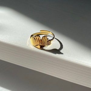 May include: A gold-colored ring with a fan-shaped design. The ring has an adjustable band and is set against a white background with shadows. The fan design is detailed and catches the light, creating a bright, reflective surface.